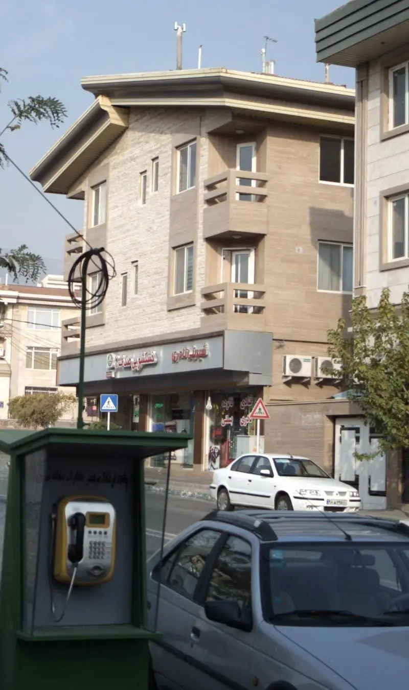 تصویر نمای خارجی قنادی شیرینی نادری در یک روز آفتابی، با ساختمان سنگی و تلفن عمومی زرد رنگ در جلوی مغازه.