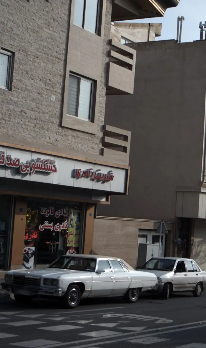 تصویر یک فروشگاه شیرینی نادری و قنادی در خیابان، با تابلوی خشکشویی صدق و نمای سنگی ساختمان و دو خودروی پارک شده.