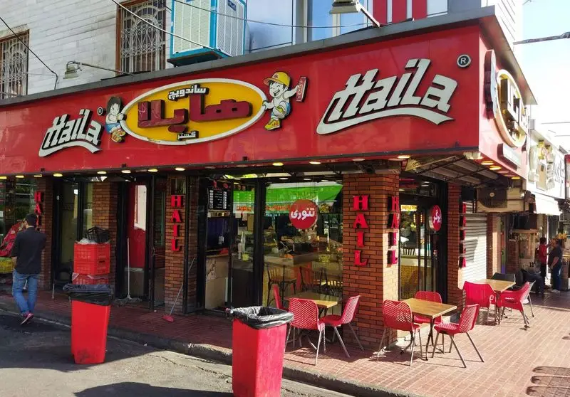 یک رستوران فست فود به نام ساندویچ هایلا با نمای قرمز برجسته، لوگوی هایلا، میز و صندلی‌های بیرون در خیابان.