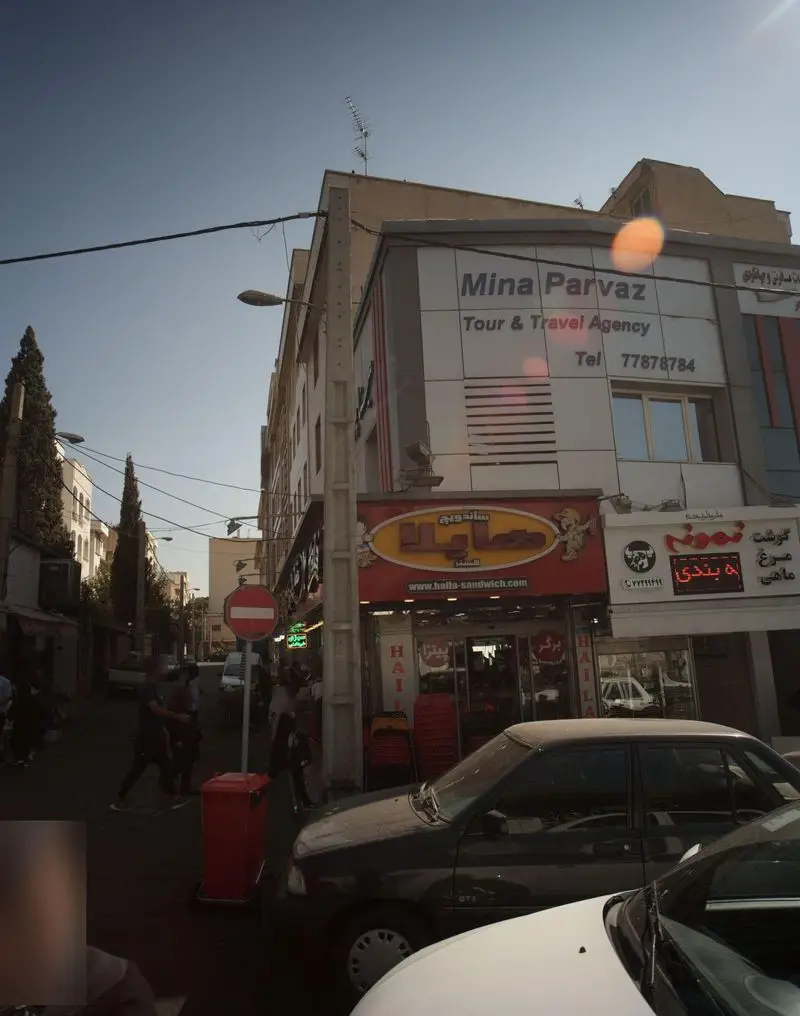 تصویر نمای بیرونی ساندویچ هایلا در خیابانی شلوغ در تهران، با تابلوی فست فود و آژانس مسافرتی مینا پرواز بالای آن.