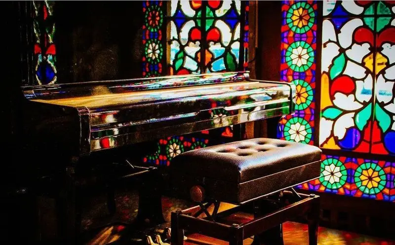 پیانوی باشکوه در کنار پنجره‌های رنگی چشم‌نواز، نشان‌دهنده دکوراسیون مجلل کاخ رستوران نقش جهان و فضایی هنری.