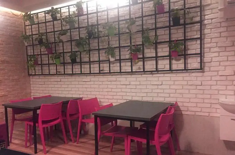 فضای داخلی رستوران ایتالیایی بولونیز: دیوارهای آجری، گلدان‌های آویزان روی شبکه مشکی و میز و صندلی‌های مدرن صورتی رنگ.