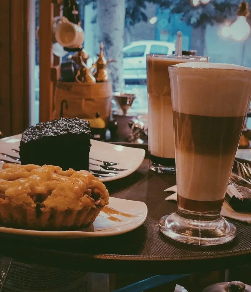 لذت چشیدن کیک شکلاتی و تارت سیب در کنار دو فنجان لاته خوش‌طعم در فضای گرم و دلپذیر کافه ایستگاه لاله.