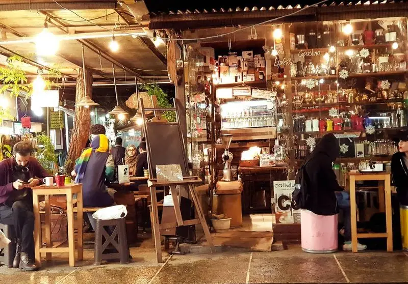 تصویری از نمای شب کافه ایستگاه لاله، با مشتریان نشسته در فضای باز و داخلی، نورپردازی گرم و انواع نوشیدنی‌های قهوه.