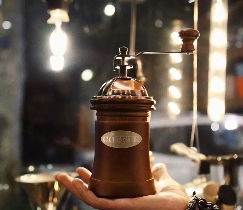 آسیاب قهوه دستی آنتیک با نشان COFFEE در دست، نمادی از تجربه اصیل قهوه تازه و عطر دلنشین در فضای گرم و دنج کافه ایستگاه لاله.