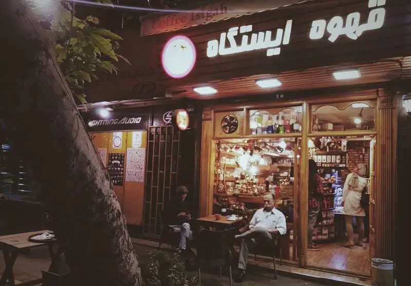 تصویری گرم و دعوت‌کننده از کافه ایستگاه لاله در شب. ویترین پر از انواع لوازم و محصولات قهوه. مشتریان در حال لذت بردن از فضای دنج و نوشیدنی‌های گرم.