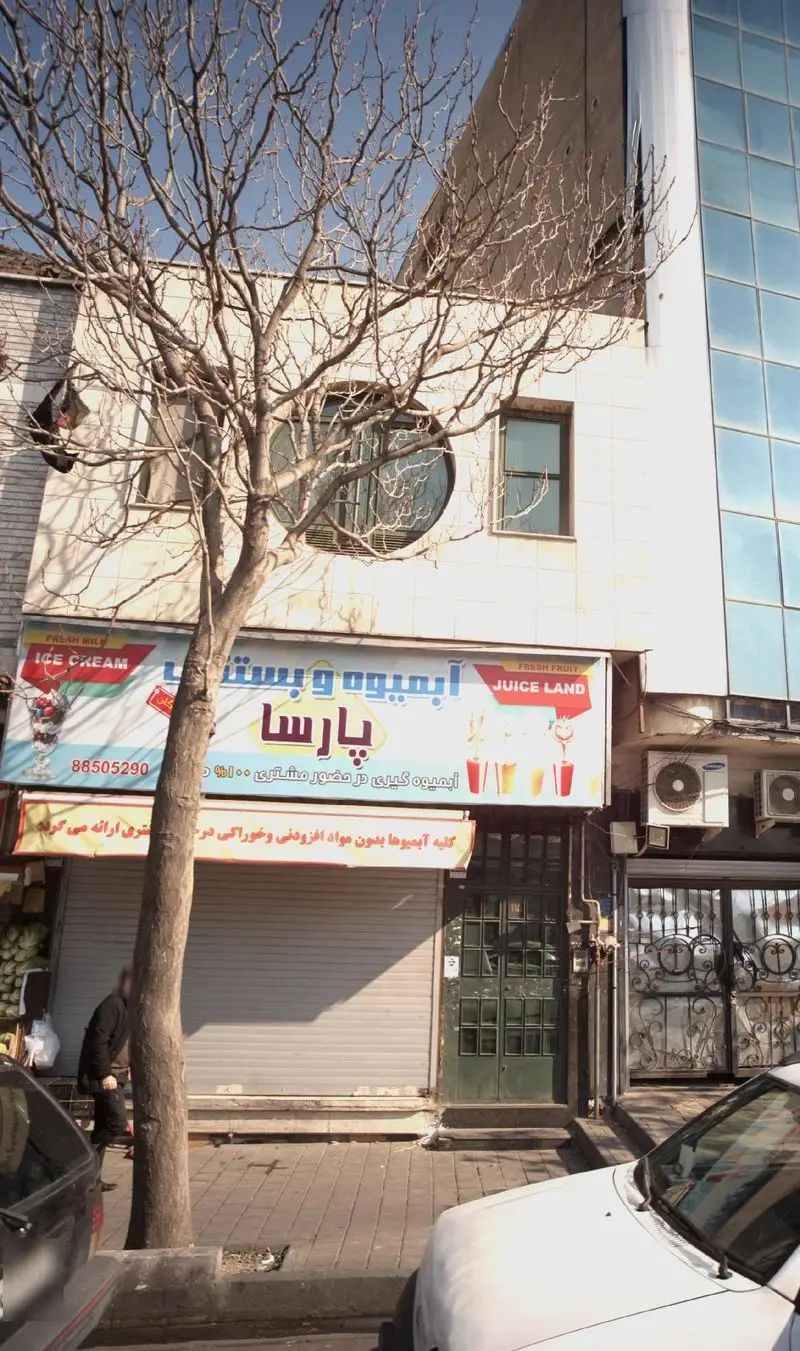 نمای بیرونی فروشگاه آبمیوه بستنی پارسا با تابلوهای "ICE CREAM" و "JUICE LAND" در خیابان، با درختی بدون برگ و ماشین‌ها.