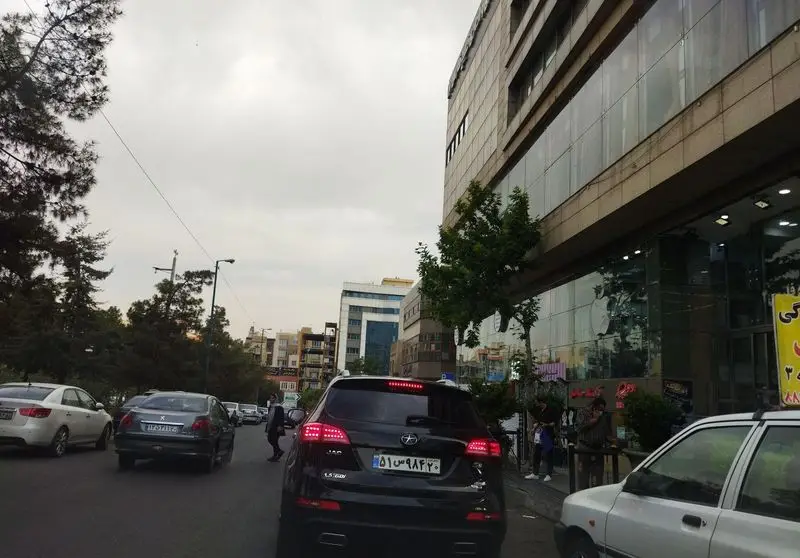 خیابان شلوغ شهری با ماشین‌ها و ساختمان‌های بلند شیشه‌ای، احتمالاً در نزدیکی رستوران شهربانو، مکانی عالی برای تجربه غذا.