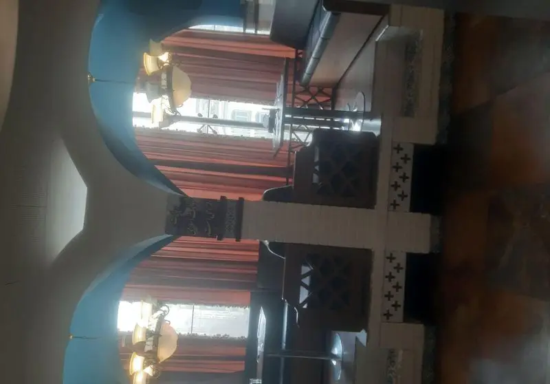 فضای داخلی زیبا و دلنشین رستوران شهربانو: سقف‌های قوس‌دار آبی و کرم، ستون‌های تزئینی، لوسترهای کلاسیک، و میز و صندلی‌های شیک کنار پنجره با پرده‌های خاص.