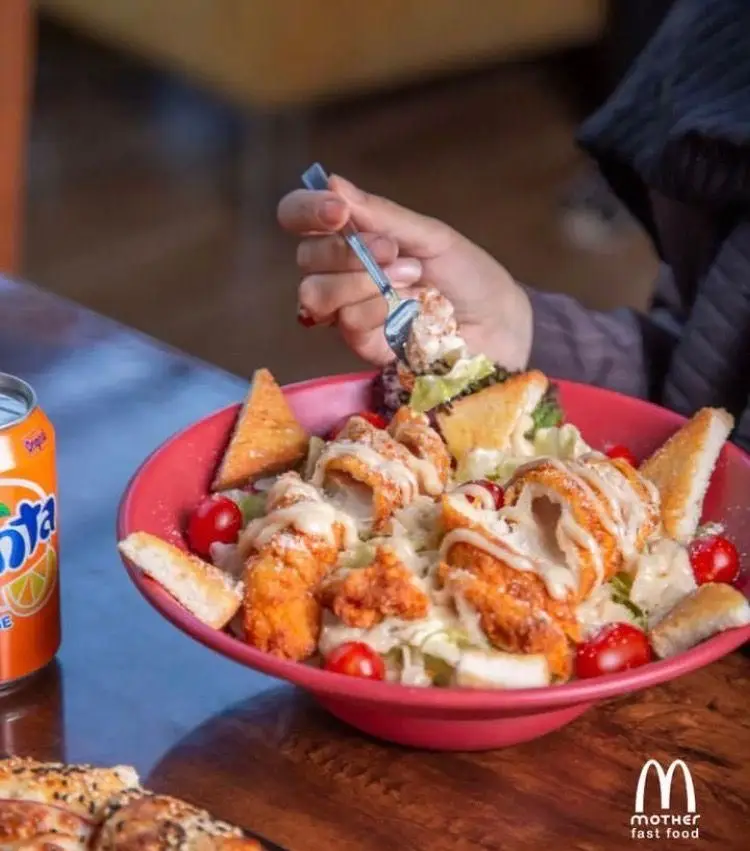 فردی در حال لذت بردن از سالاد سزار با مرغ سوخاری، گوجه گیلاسی و نان تست همراه با نوشابه فانتا از فست فود مادر.