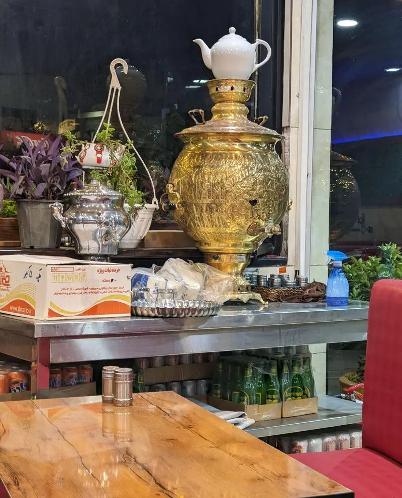 نمای داخلی جگرسرای خندان با سماور سنتی طلایی و قوری چای، سماور نقره‌ای، گلدان‌ها، میز چوبی و انواع نوشیدنی‌ها برای مشتریان.