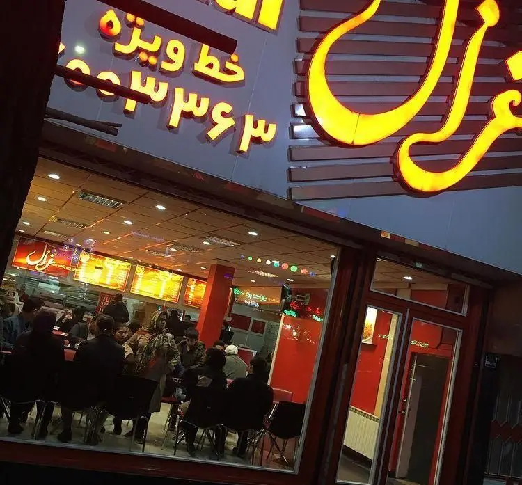 تصویری از نمای شب پیتزا غزال، تابلوی نورانی غزال و خط ویژه سفارش ۰۰۳۶۳۴۶۳، با مشتریان در فضای داخلی شلوغ رستوران.