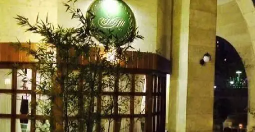 نمای شبانه فست فود بیزی کافه با تابلوی سبز براق، ورودی چوبی شیک و گیاهان بامبو. فضایی آرامش‌بخش و دلپذیر برای تجربه فست فود.