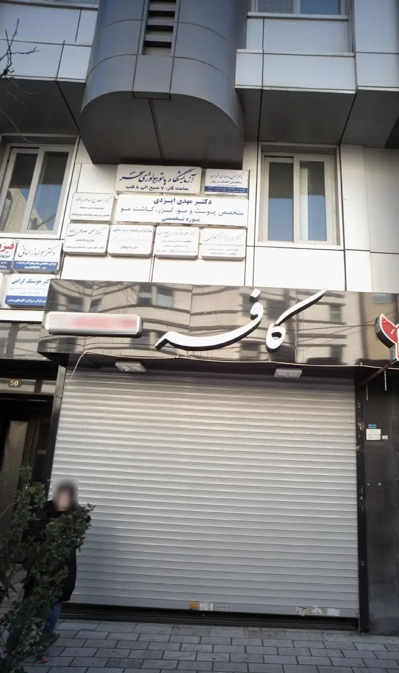 کافه با تابلوی برجسته سفید و کرکره بسته، همراه با تابلوهای مختلف پزشکان و آزمایشگاه در بالای ساختمان تجاری در شهر.