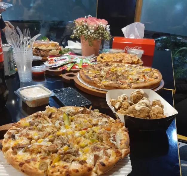 میز پر از غذاهای لذیذ فست فود بلوط شامل دو پیتزای بزرگ، برگر و مرغ سوخاری، آماده سرو در محیطی دلنشین و دوستانه