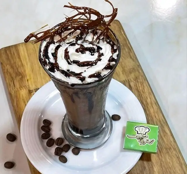 میلک شیک شکلاتی لوکس با خامه، سس و دکور شکلات روی میز چوبی، از نوشیدنی های خاص باغ رستوران طبیعت برای تجربه ای متفاوت.