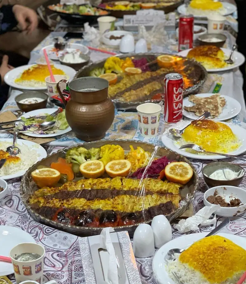 سفره رنگین و پر از غذاهای لذیذ ایرانی شامل انواع کباب، برنج زعفرانی و ماهی در باغ رستوران طبیعت، مناسب دورهمی خانوادگی.