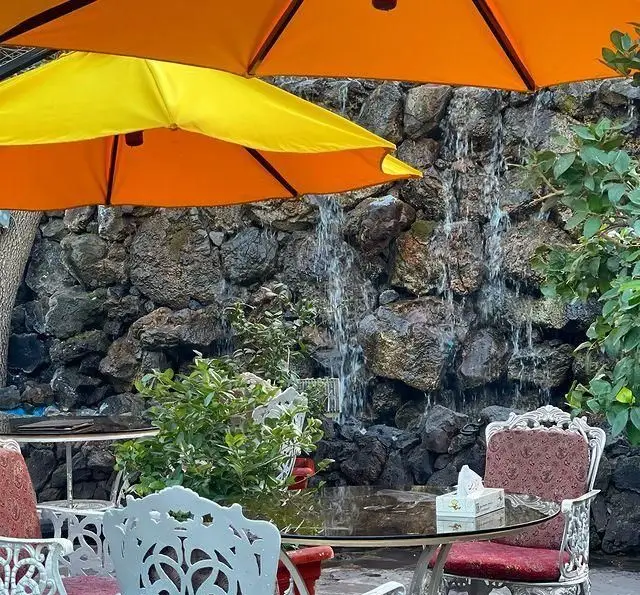 فضای باز دلنشین باغ رستوران طبیعت با آبشار سنگی زیبا، چترهای آفتاب‌گیر و میز و صندلی‌های مجلل مناسب برای تجربه غذایی لذت‌بخش.