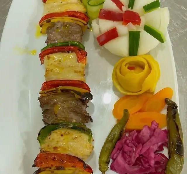 کباب میکس گوشت و سبزیجات متنوع با پنیر در بشقاب سفید، به همراه گارنیش شیک پیاز، هویج، کلم و فلفل در باغ رستوران طبیعت.