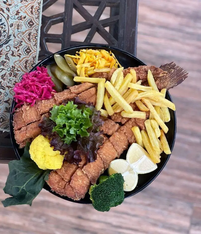سینی بزرگ ماهی سوخاری کامل، شنیسل ماهی، سیب زمینی سرخ کرده فراوان، و دورچین رنگارنگ از ترشی و سبزیجات در باغ رستوران طبیعت.