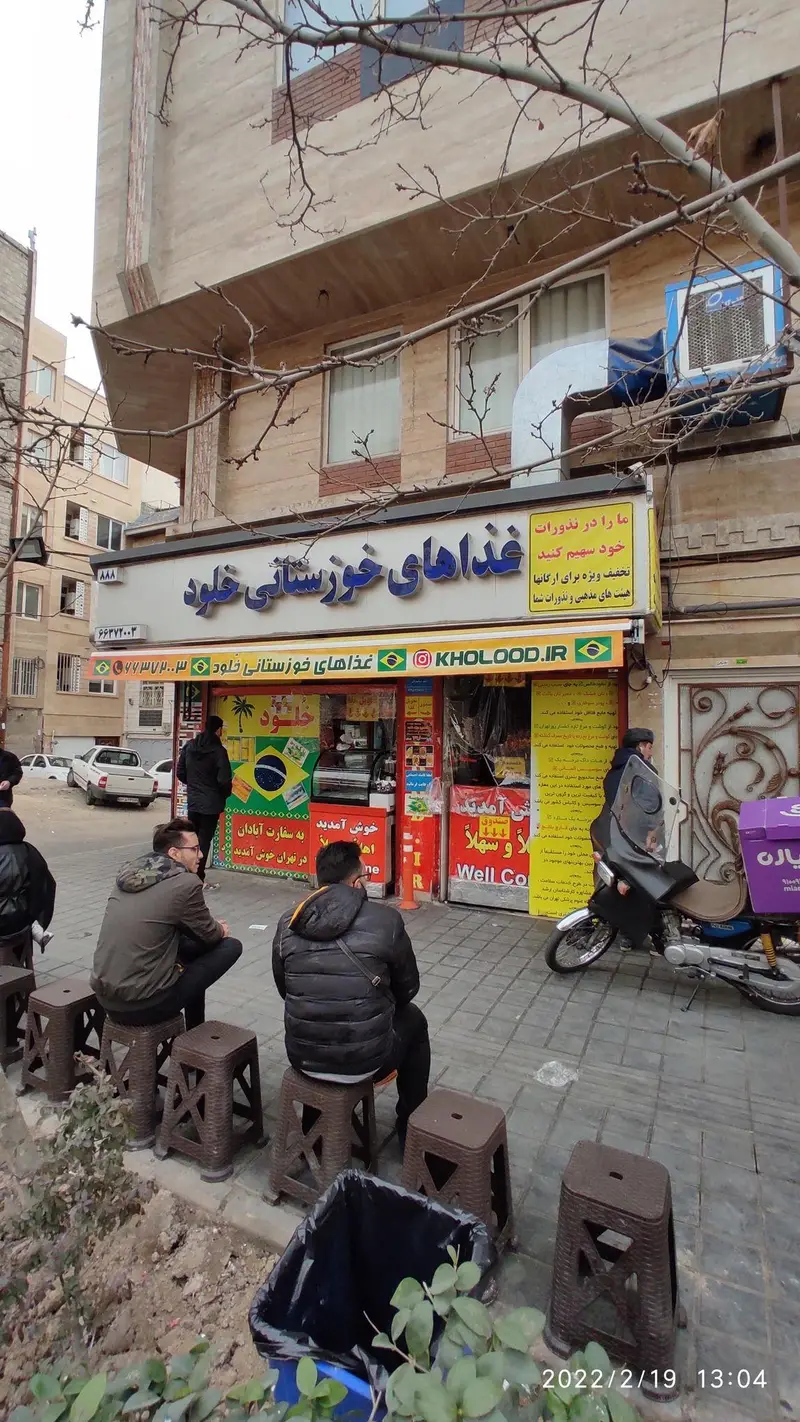 نمای بیرونی ساندویچی خلود، رستوران غذاهای خوزستانی در تهران، با مشتریان در حال انتظار و موتور سیکلت بیرون‌بر. فضای شلوغ خیابان.