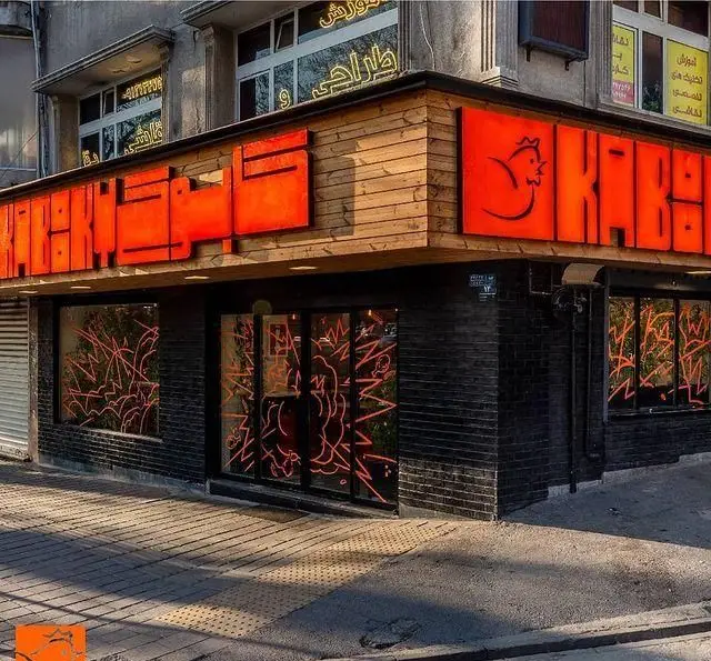 تصویر جذاب از نمای بیرونی مرغ سوخاری کابوکی با تابلوی نئونی نارنجی، دیوارهای آجری تیره و گرافیک‌های روی شیشه.