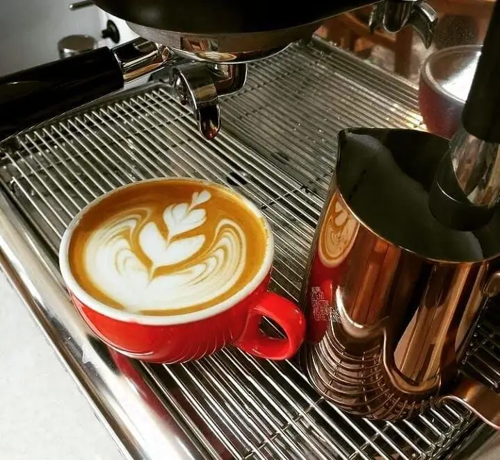 قهوه لاته آرت هنرمندانه در فنجان قرمز رنگ، آماده سرو در کافه قهوه ساوا. نمای نزدیک از دستگاه اسپرسو و شیرجوش براق.