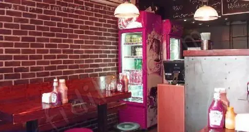 فضای داخلی دکان برگر با دیوار آجری، میزهای چوبی قرمز و نیمکت‌ها، یخچال‌های صورتی نوشیدنی و پیشخوان فلزی. محیطی مدرن و دلنشین برای لذت بردن از همبرگر.