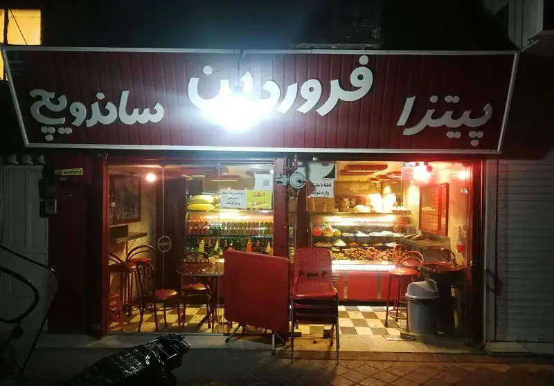 نمای شب فست فود پیتزا فروردین با تابلو روشن پیتزا و ساندویچ، ویترین‌های پر از خوراکی و میز و صندلی داخل مغازه