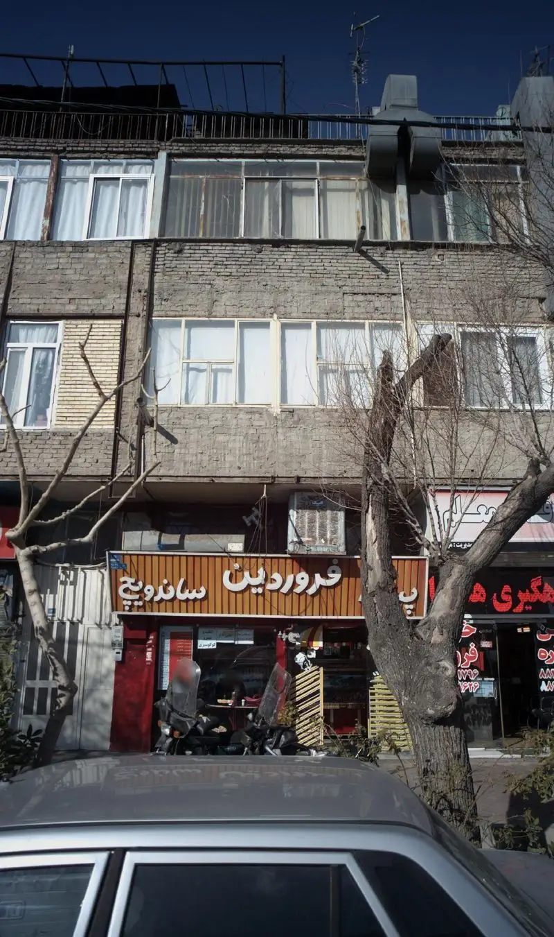 نمای بیرونی پیتزا و ساندویچ فروردین در یک ساختمان شهری. تابلوی روشن فروردین ساندویچ، طبقات مسکونی و درختان خیابان.