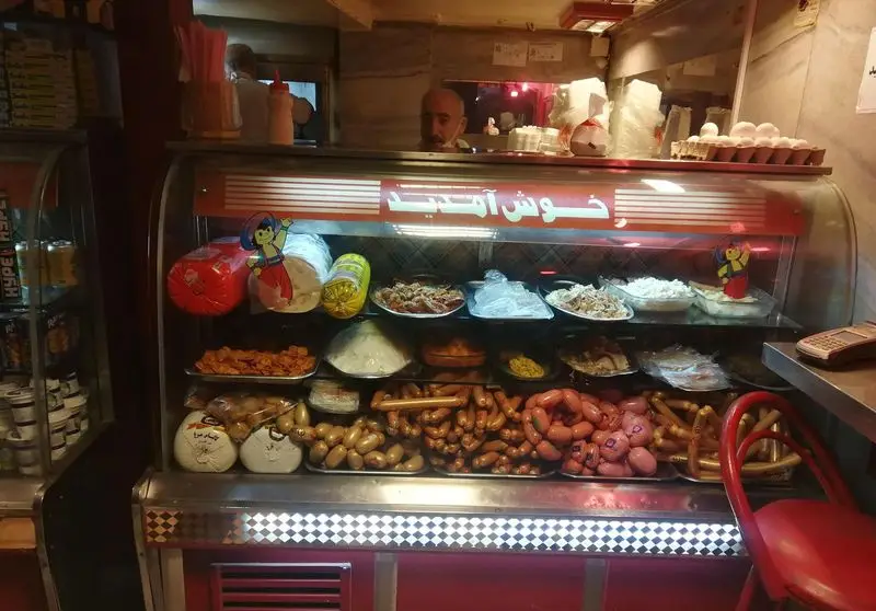 نمایی نزدیک از ویترین اغذیه پیتزا فروردین، نمایش دهنده مجموعه‌ای متنوع از سوسیس، کالباس، سالادها و مواد غذایی تازه و آماده.