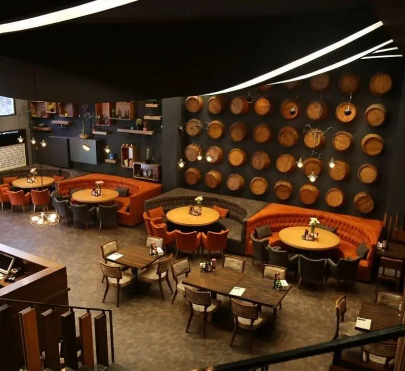 تصویری از فضای داخلی بزرگ و زیبای رستوران پیتزا میخوش با دکوراسیون مدرن، میز و صندلی‌های چوبی و مبلمان نارنجی و خاکستری، محیطی عالی برای غذاخوری.