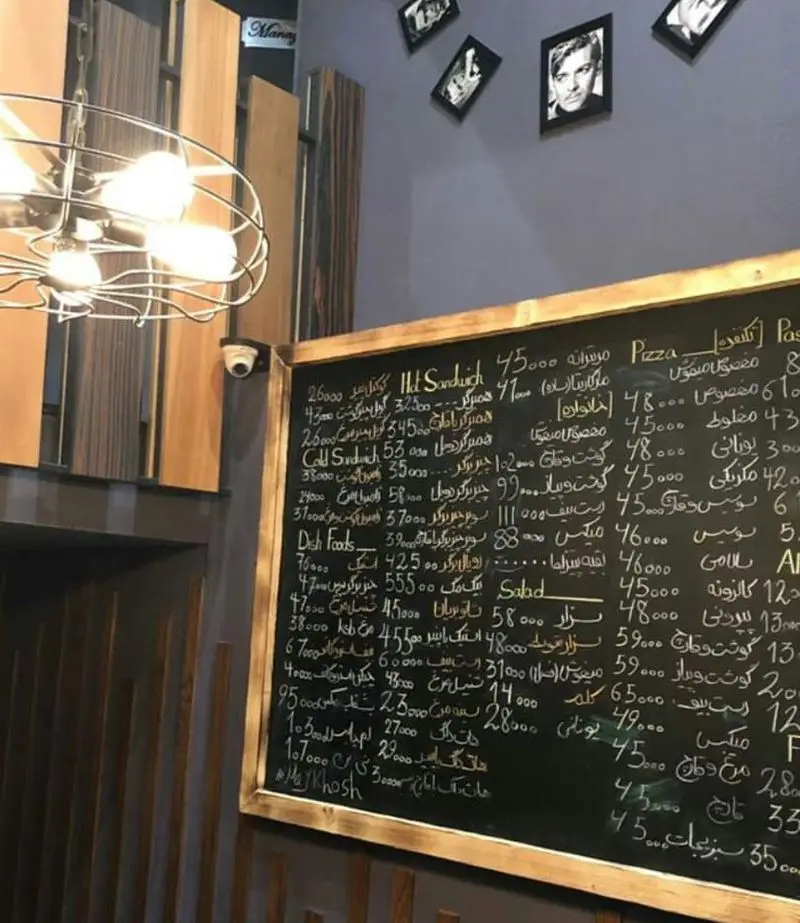 تصویر منوی پیتزا میخوش: لیست قیمت‌ها و انواع پیتزا، ساندویچ، سالاد و غذاهای رستوران روی تخته سیاه در فضایی گرم و مدرن.