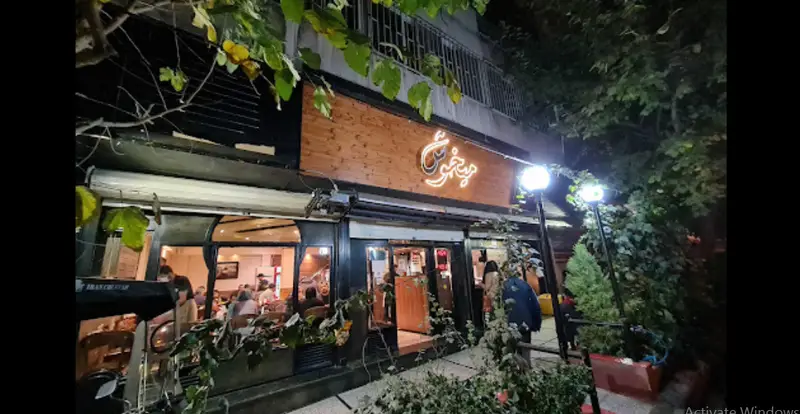 نمای شبانه و جذاب از رستوران پیتزا میخوش، با تابلوی نئونی روشن و دکور چوبی مدرن، مشتریان در حال لذت بردن از غذا در فضای داخلی، و گیاهان سبز جلوی ورودی.