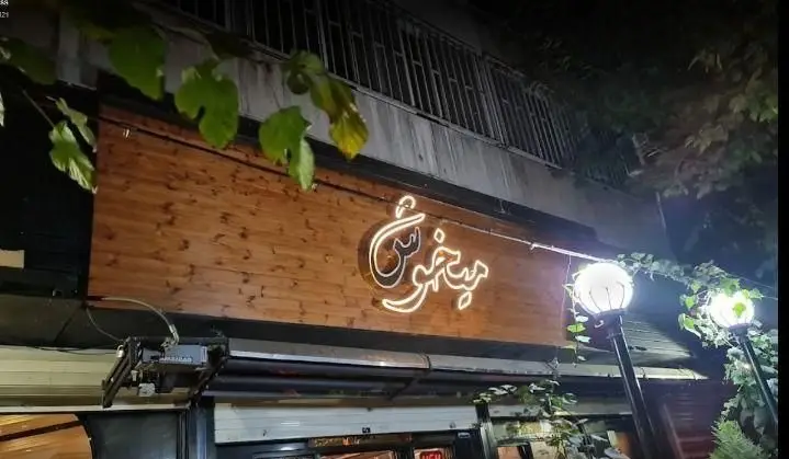 تصویری زیبا در شب از تابلوی نئونی 'پیتزا میخوش' روی سردر چوبی رستوران، با نورپردازی گرم و برگ‌های درخت انگور.