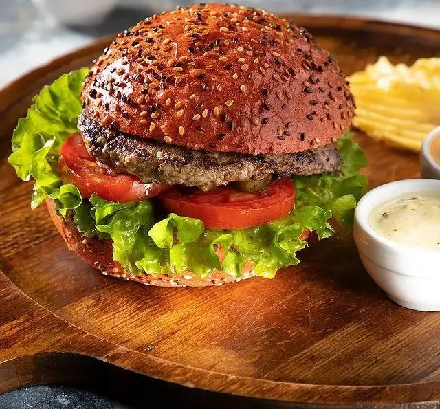 همبرگر لذیذ و اشتهاآور کافه رستوران مگریز با نان ویژه کنجدی، گوشت آبدار، گوجه و کاهوی تازه، سرو شده روی تخته چوبی.