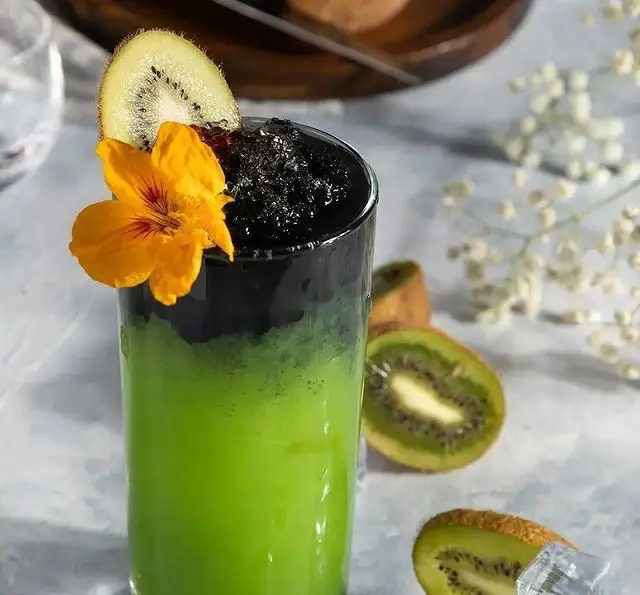 نوشیدنی خنک و رنگی کافه رستوران مگریز با طعم کیوی و لایه مشکی خاص، تزیین شده با گل و کیوی، مناسب برای تابستان و لحظات دلپذیر.