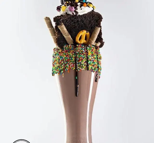 تصویر شیک شکلاتی دست‌ساز و خاص کافه رستوران مگریز با تزئین برانی، خامه، اسپرینکل‌های رنگی و چوب شور.