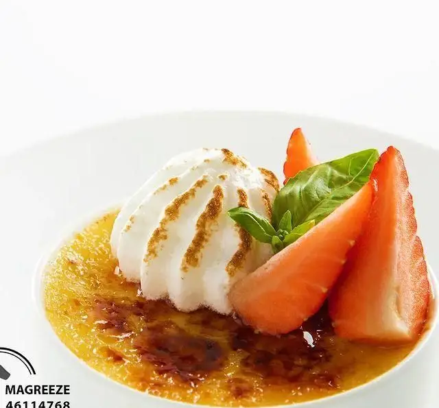 دسر کرم بروله لذیذ کافه رستوران مگریز با خامه فرم گرفته، توت فرنگی تازه برش خورده و برگ معطر ریحان، تصویری اشتهاآور.