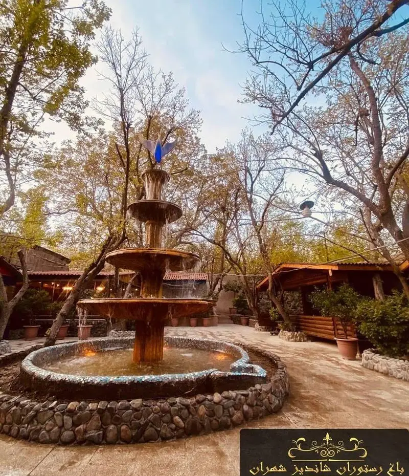 نمایی زیبا از باغ رستوران شاندیز تهران با فواره‌ای چشم‌نواز، آلاچیق‌های چوبی و درختان سرسبز، محیطی دلنشین برای صرف غذا.