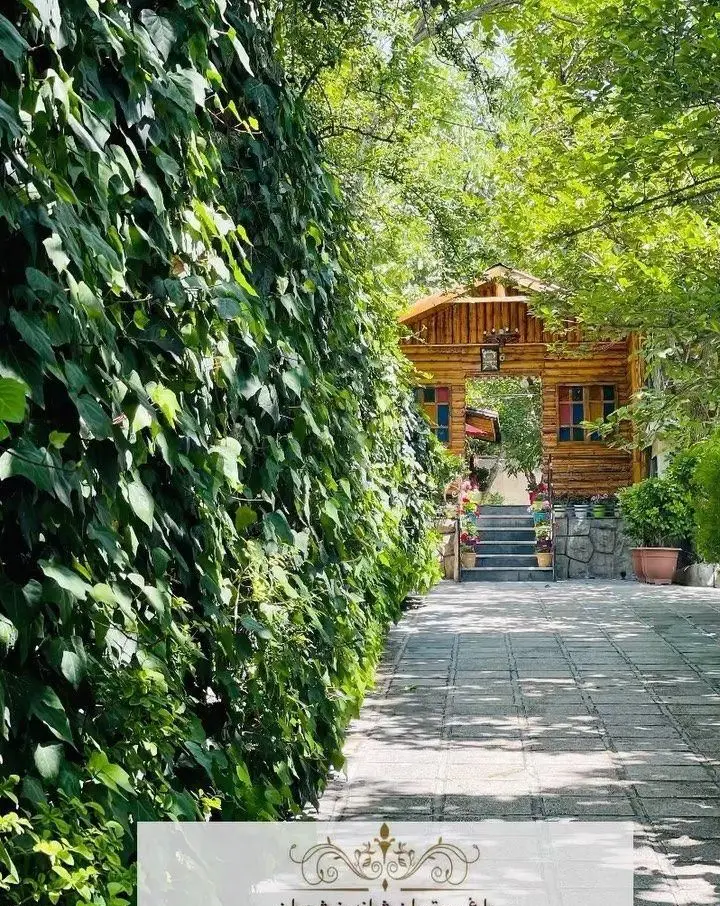 باغ رستوران شاندیز تهران: ورودی دلنشین با دیوار سبز انبوه از پیچک، مسیر سنگفرش، و سازه چوبی زیبا در محیطی سرسبز و طبیعی