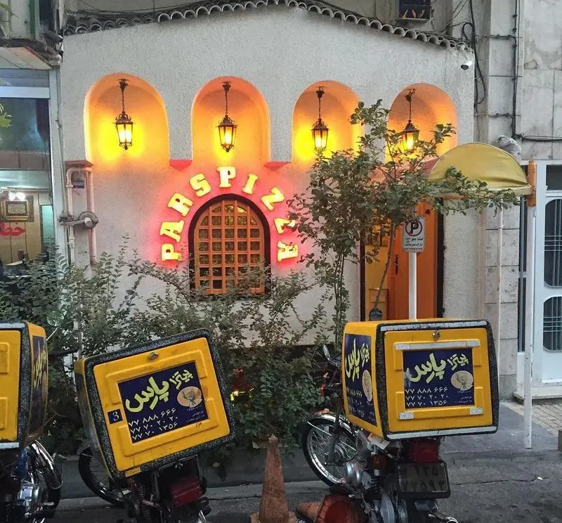 پیتزا پارس، نمای جذاب بیرونی رستوران فست فود با تابلو نئون درخشان و موتورهای زردرنگ پیک برای تحویل سریع سفارش.