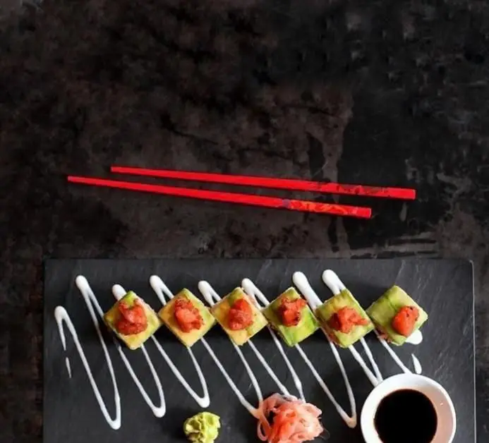 سوشی رول‌های خوشمزه آووکادو و تن ماهی رستوران واسابی، سرو شده روی بشقاب سنگی تیره با سس و چوب چاپستیک قرمز، غذای ژاپنی لذیذ.