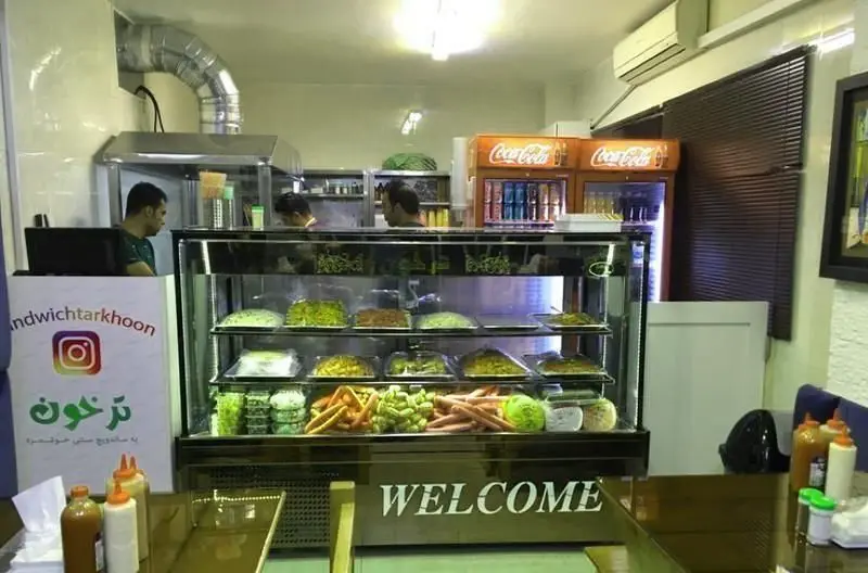 نمای داخلی کسب و کار ساندویچ ترخون؛ ویترین مدرن غذاهای آماده و تازه، نوشیدنی‌ها و پرسنل فعال، محیطی دلپذیر فست‌فود.