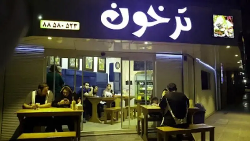نمای شبانه از ساندویچ فروشی ترخون با تابلوی روشن، مشتریان در فضای باز و داخل، همراه با شماره تماس ۸۸۵۸۰۵۲۳