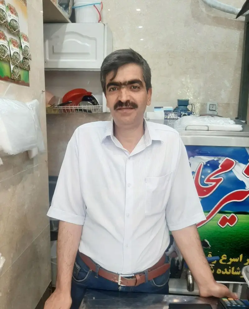 فروشنده لبنیات سنتی ناصری لیقوانی با چهره ای مهربان در مغازه خود ایستاده و آماده عرضه محصولات تازه و سنتی است.