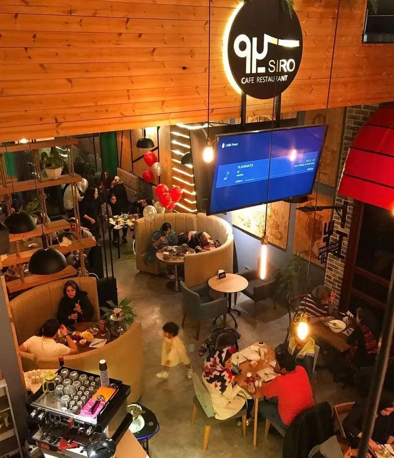 فضای داخلی دنج کافه رستوران سیرو با مشتریان در حال صرف غذا و نوشیدنی. دکوراسیون مدرن، سقف چوبی و محیط گرم مناسب دورهمی و ملاقات‌ها.