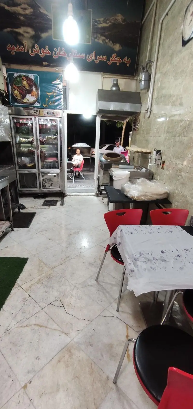 نمای داخلی جگرسرای ستارخان، یک رستوران کبابی با فضای گرم و صمیمی، شامل میز و صندلی، یخچال غذا و تنور نان در حال کار.