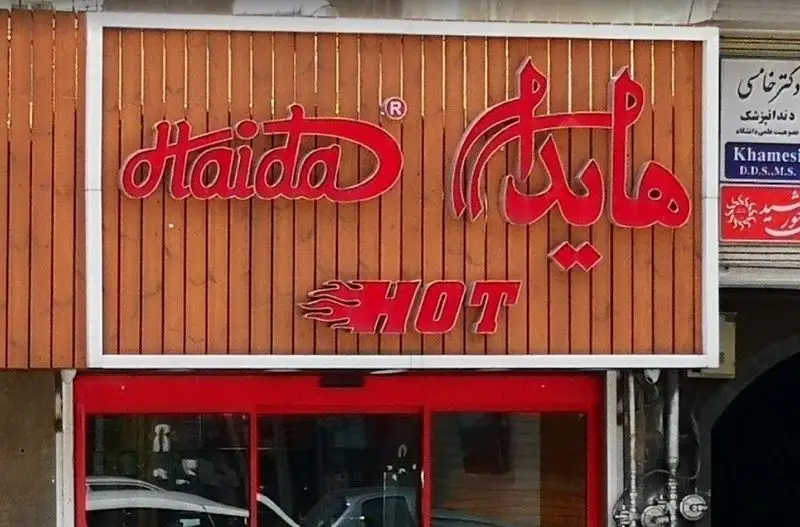 نمای بیرونی شعبه‌ای از ساندویچ هایدا (Haida)، با تابلوی قرمز رنگ برجسته شامل نام فارسی، انگلیسی و "HOT" با لوگو شعله روی پس‌زمینه چوبی.