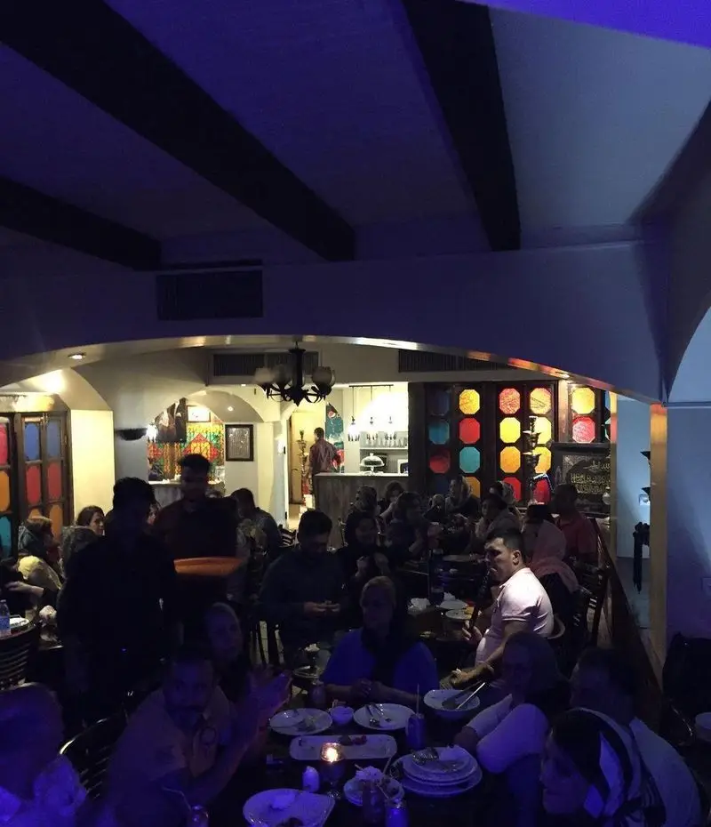 فضای داخلی رستوران سنتی عمارت با مشتریان در حال صرف غذا. دکوراسیون سنتی ایرانی، پنجره‌های رنگی و نورپردازی آرامش‌بخش برای تجربه‌ای دلنشین.